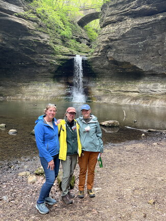 Three women pose for a photo in front of a waterfall with a bridge on the background