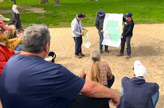 A group of people watch a man explain something on a map while two others hold up the map