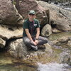Jenna Shelton. A woman sitting on a rock next to a creek posing for the camera.