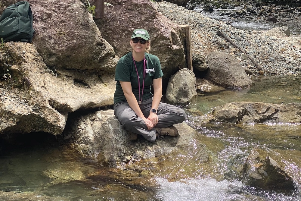Jenna Shelton. A woman sitting on a rock next to a creek posing for the camera.